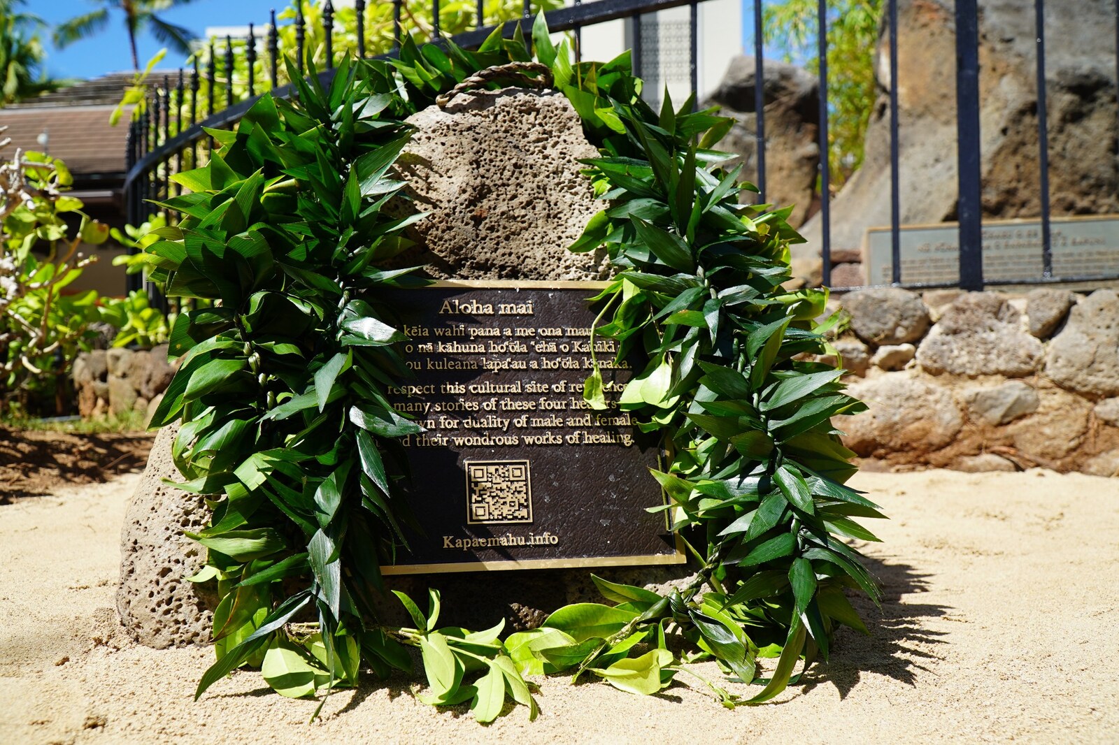 A lei-covered Kapāemahu plaque and QR code in front of the healing stones at Kūhiō Beach Park.