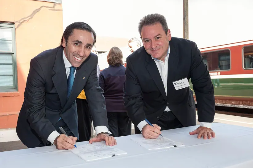 Two men in dark suits stand leaning over a table covered with a white cloth, each signing a document with a pen and smiling toward the camera; other people stand behind them and a train is visible at the right.